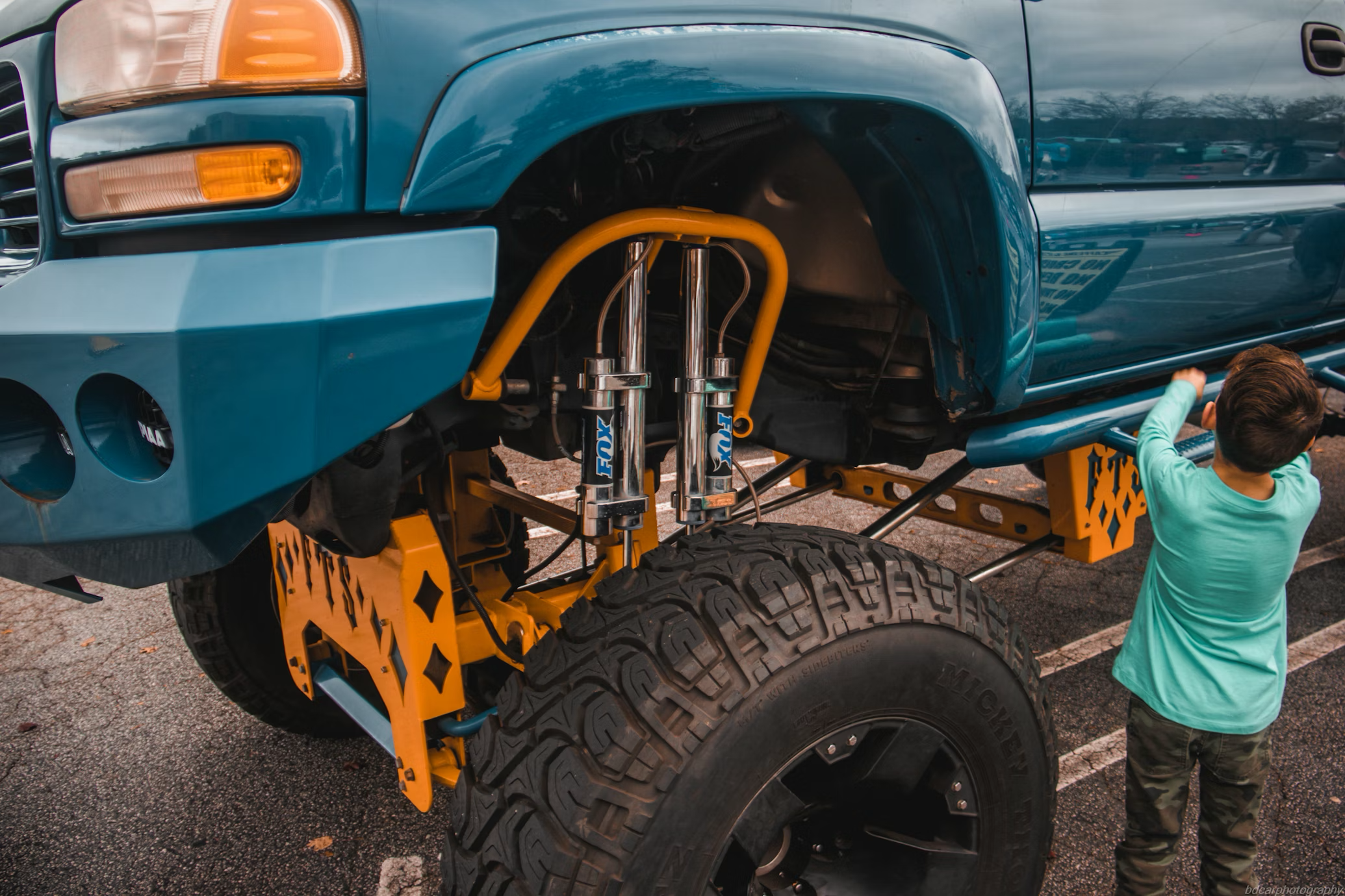 A highly lifted teal truck with a large yellow suspension lift kit, FOX shocks, and a child looking at the side step